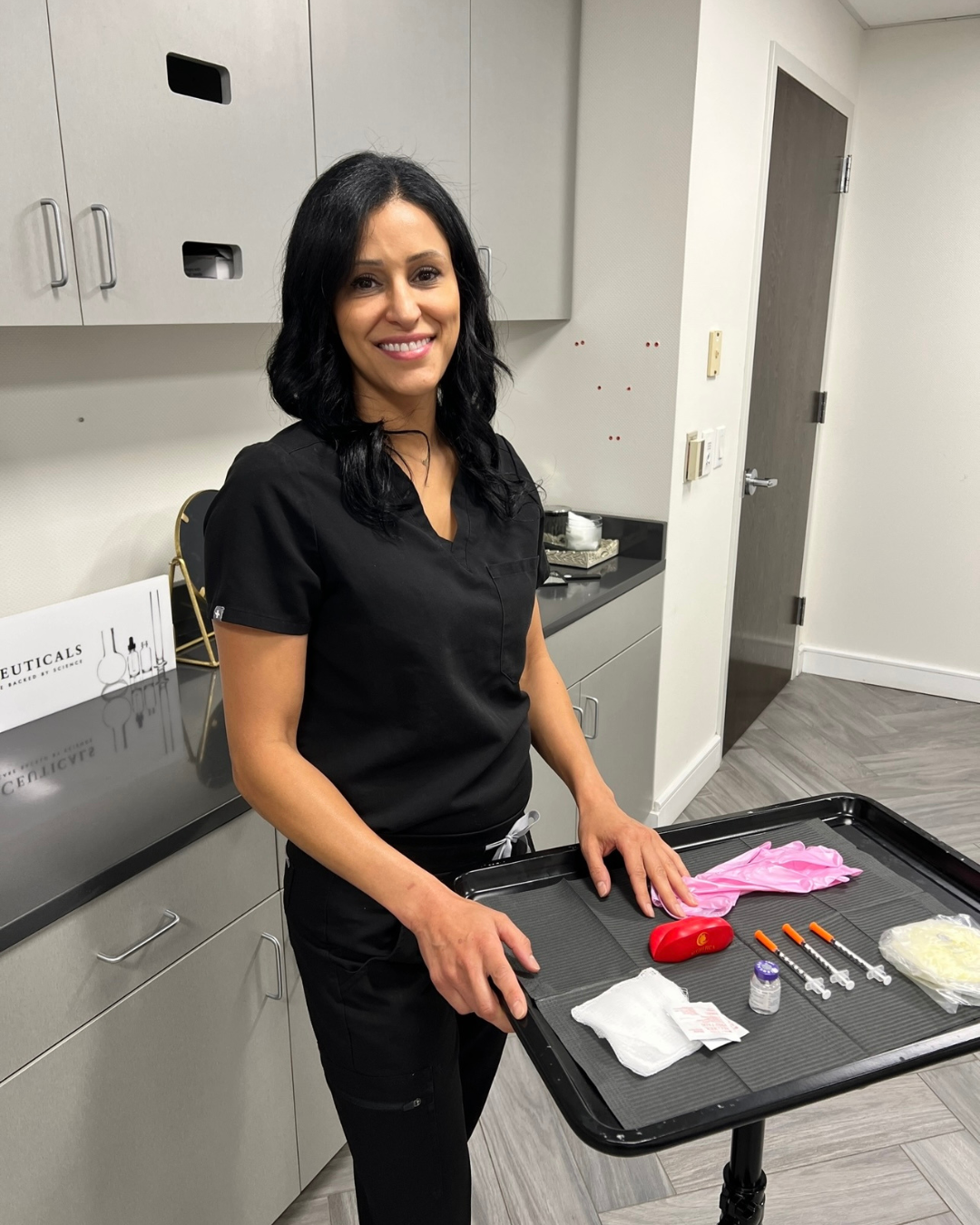 Smiling healthcare professional in black scrubs standing by a prep cart with medical supplies in a clinic. (informative)
