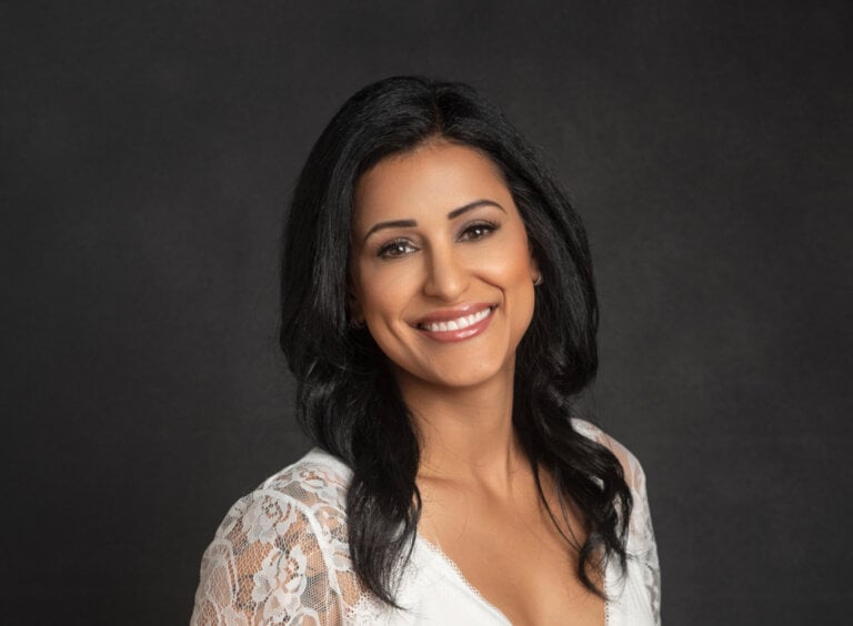 Smiling woman with shoulder-length black hair wearing a white lace top and jeans, posed against a dark studio background.
