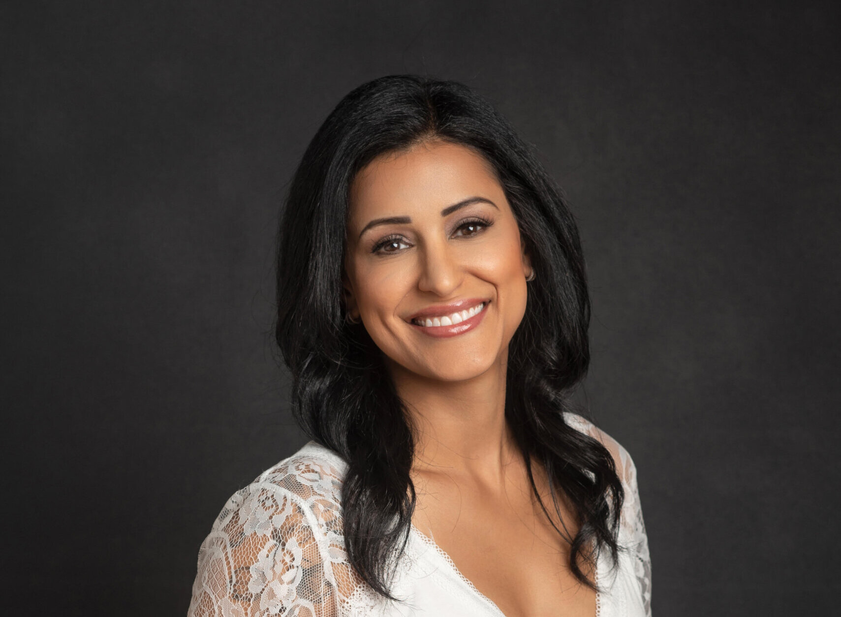 Smiling woman with shoulder-length black hair wearing a white lace top and jeans, posed against a dark studio background.