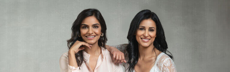 Two women sit cross-legged on a wooden floor, smiling at the camera with their arms around each other in casual outfits.