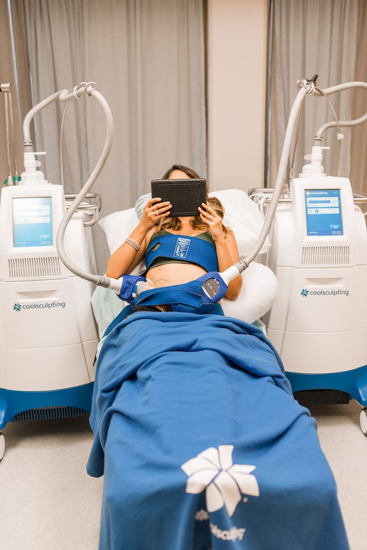 Person resting during a CoolSculpting treatment, lying in a clinic bed with blue wrap and handheld tablet.