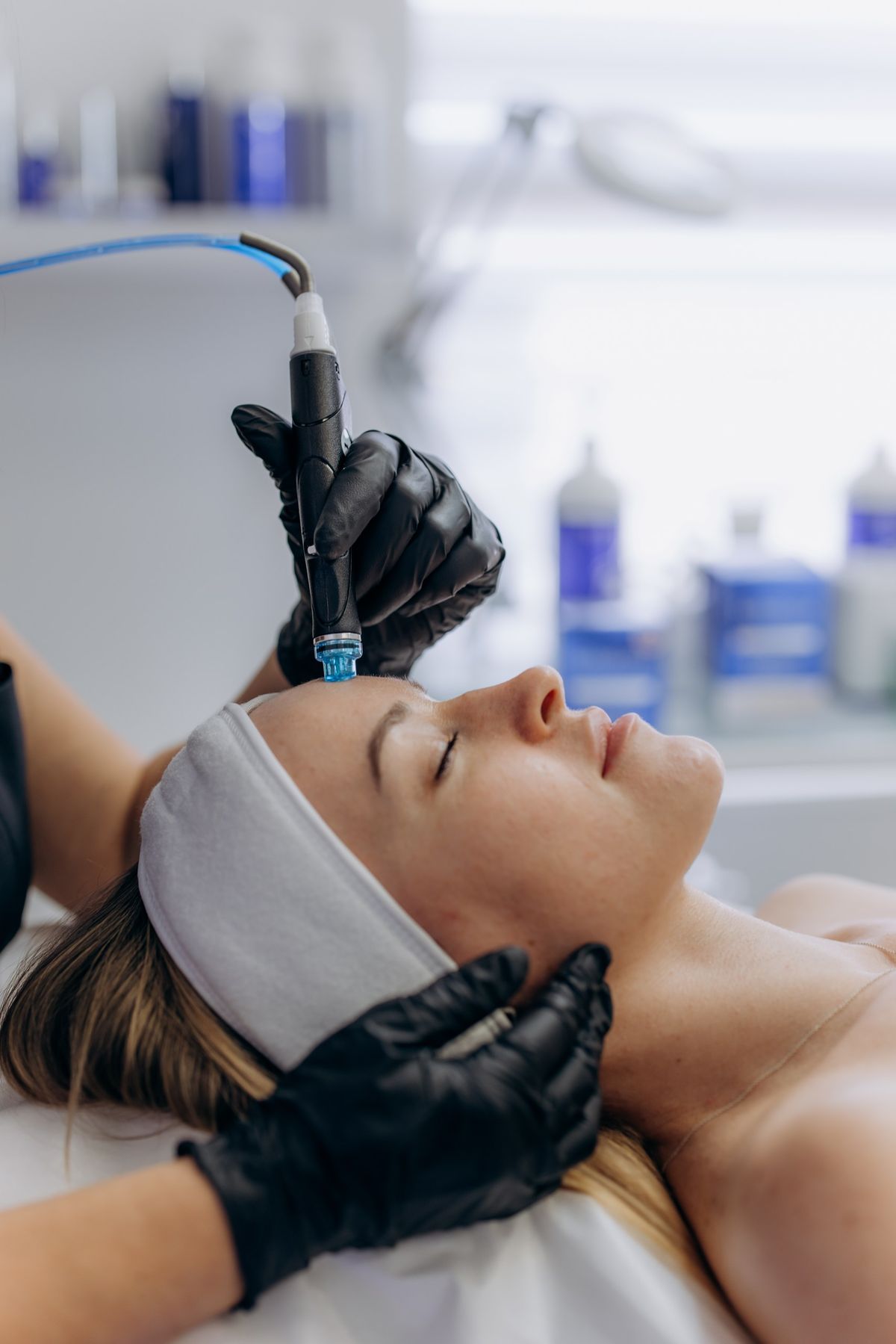 Woman receiving a cosmetic facial; gloved technician uses a handheld pen-shaped device on her forehead as she rests with a headband on.