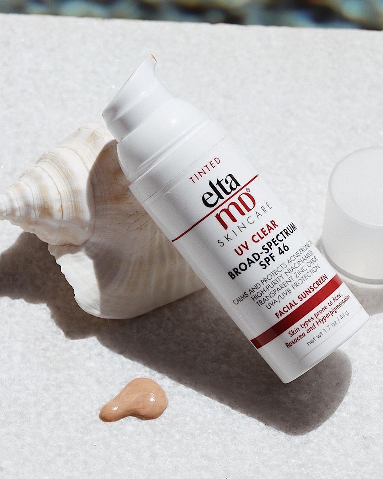 Sunscreen tube labeled EltaMD UV Clear SPF 46 on white sand beside a seashell and a dab of lotion visible nearby