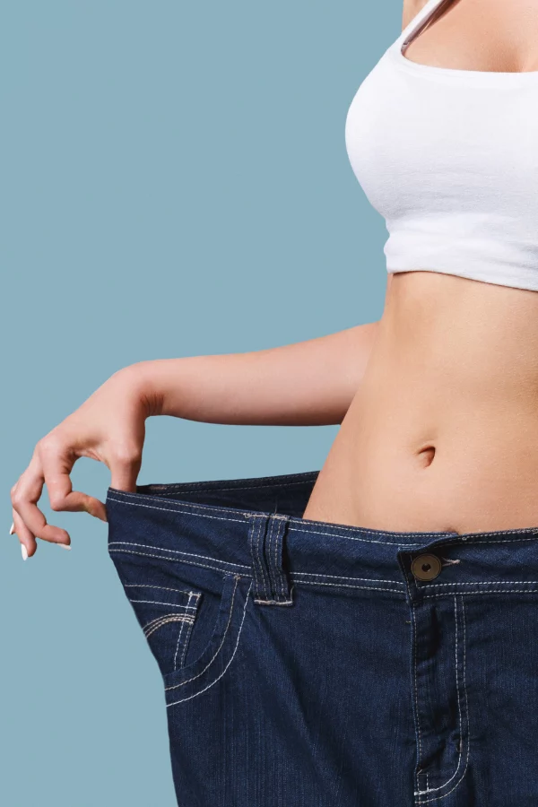 Person wearing a white crop top holds out loose-fitting jeans at the waist to show extra room in the waist area.
